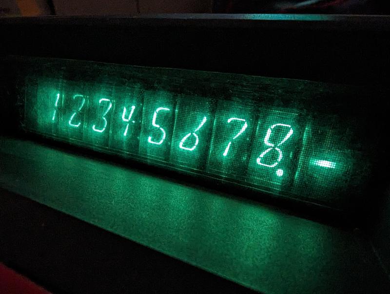 A close-up of the nine VFD tubes, showing the digits 1 to 8, followed by a decimal point and a minus sign. The environment is dark. The tubes glow a bright green tint.